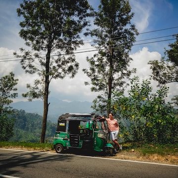 A person wearing a peach shirt and white shorts leans against a green tuk-tuk parked beside a road lined with tall trees. The background features lush greenery and distant hills under a blue sky with scattered clouds.